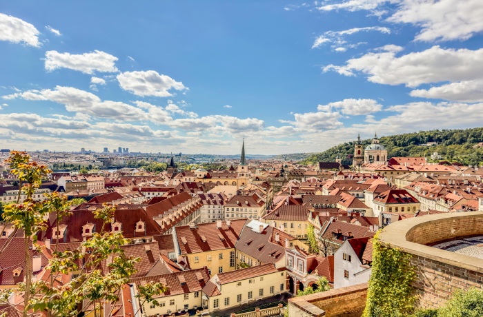 Ein Panoramablick auf Prag mit vielen roten Ziegeldächern, engen Straßen und markanten Kirchtürmen, eingebettet in eine grüne Hügellandschaft unter einem blauen Himmel mit weißen Wolken.