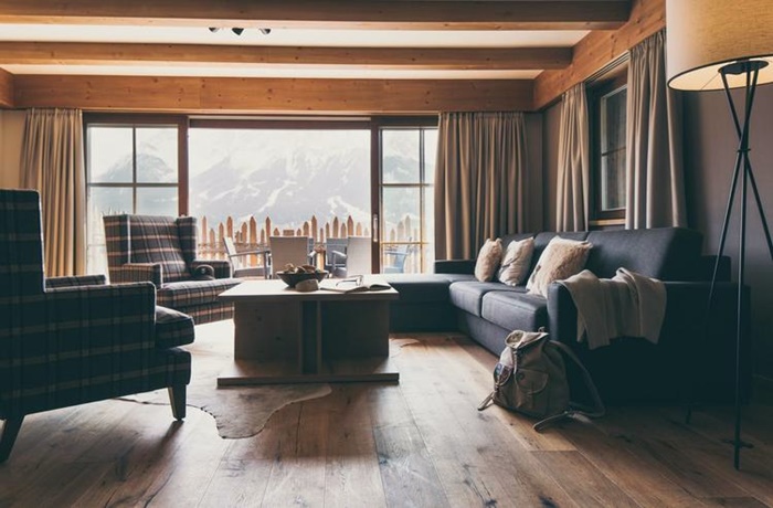 Wohnzimmer eines Chalets mit karierten Sesseln und schwarzem Sofa, Holzboden und Holzbalken, Balkon mit Ausblick auf die Berge