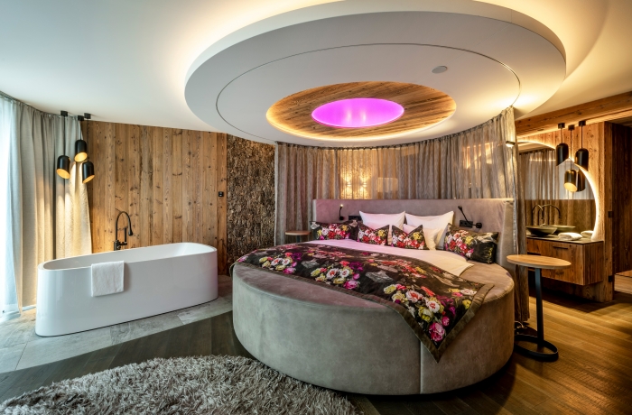 Hotelzimmer mit rundem Bett, freistehendem Whirlpool, Holzwänden und indirekter Deckenbeleuchtung.